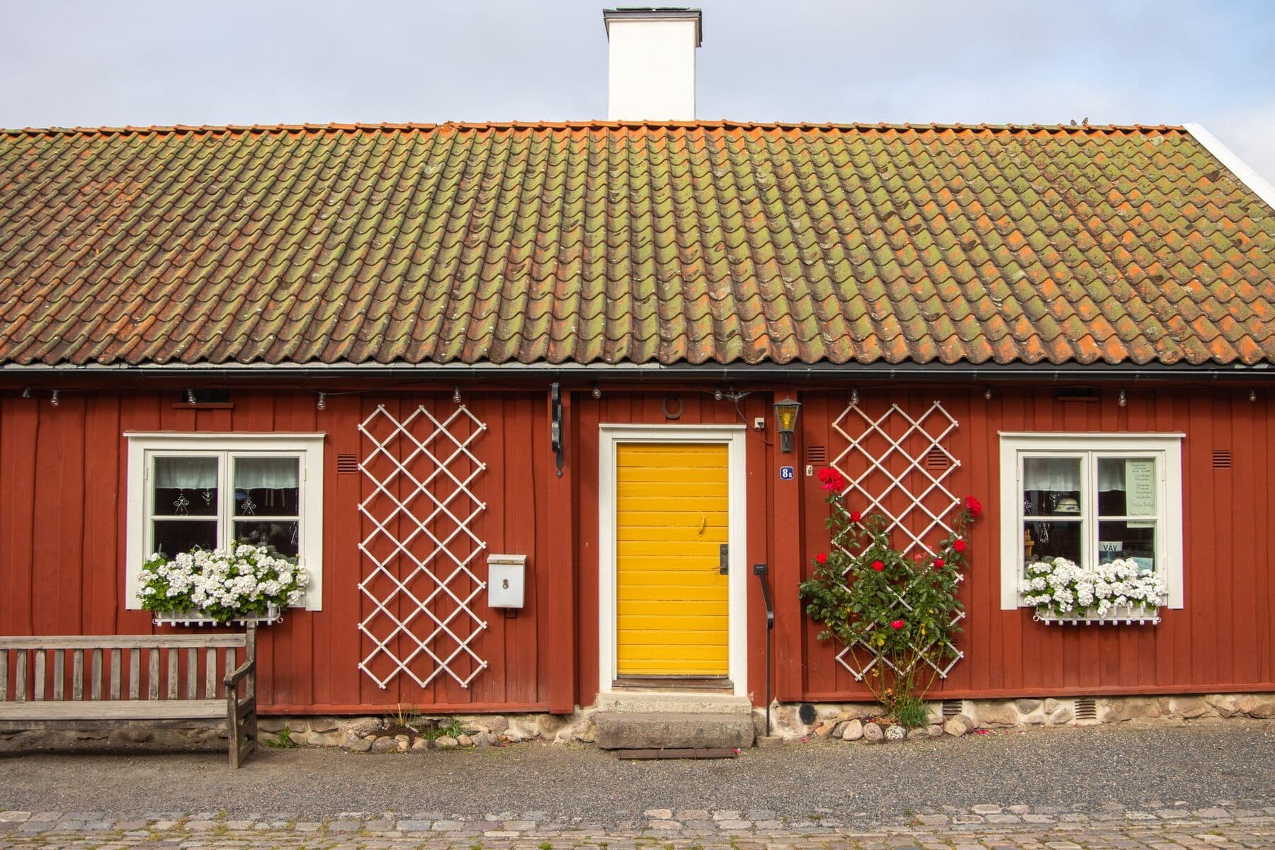 Ett rött svenskt trähus med gul dörr, vita fönster med blomlådor och en bänk framför